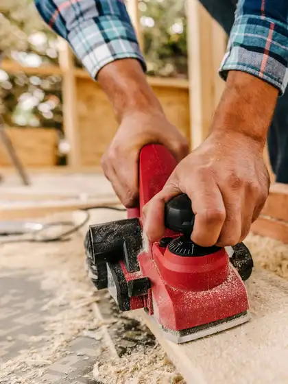 Hände am Elektrohobel, daneben Hobelspäne auf dem Holz