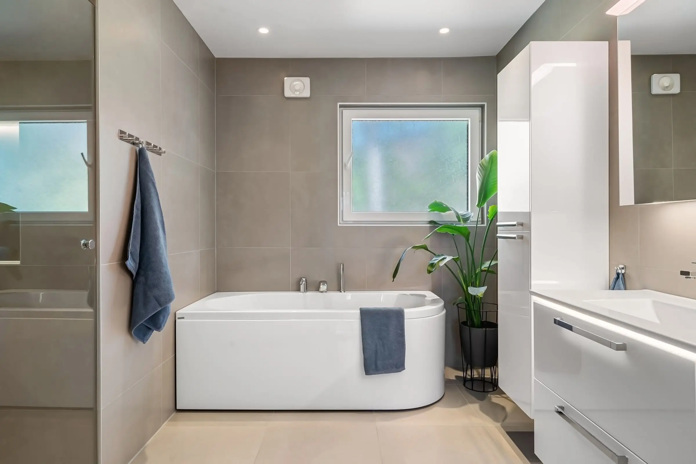 Familienbad mit Wanne unter dem Milchglasfenster, graue Großformatfliesen, rechts Hochschrank und Waschtisch