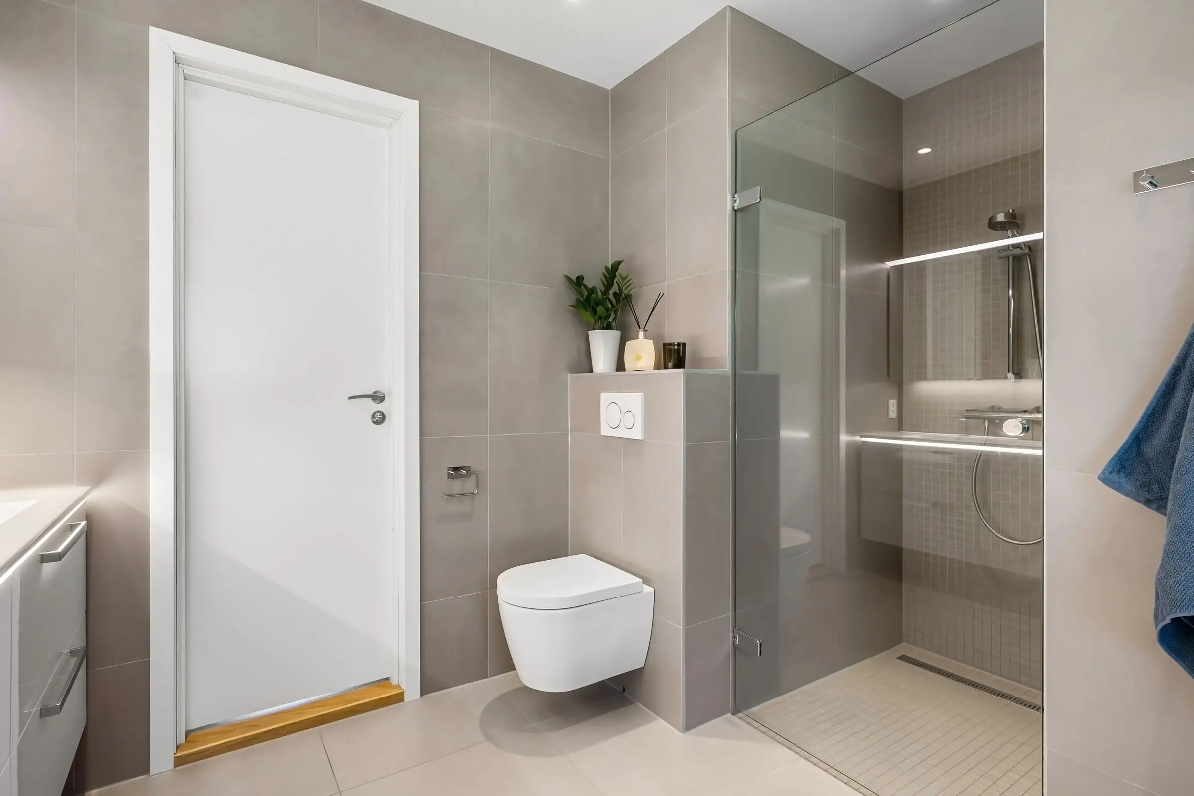 Bad mit weißer Tür, WC an einer halbhohen Vorwand und bodengleicher Dusche hinter Glas