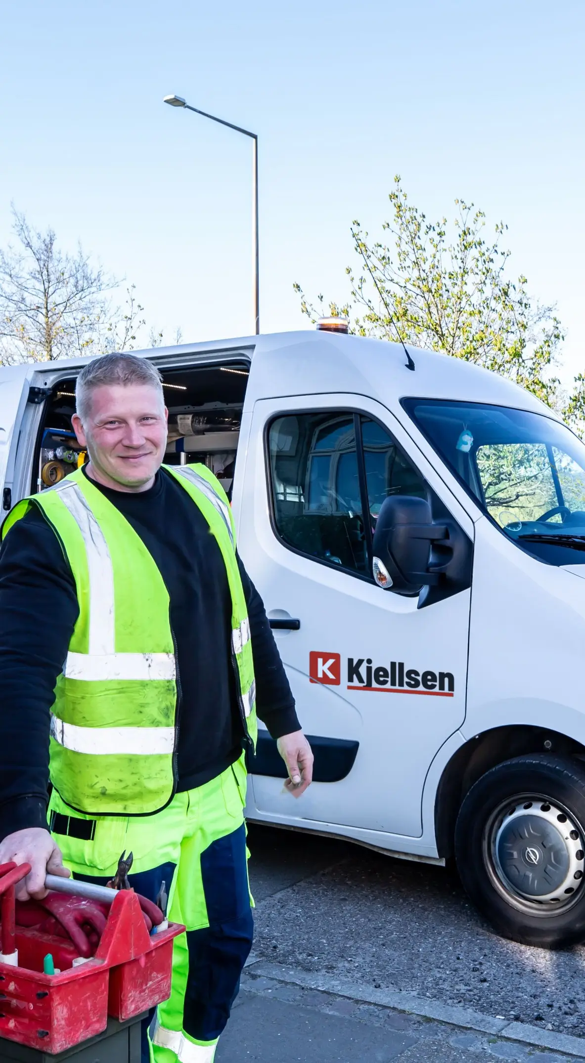 Daniel Ahrens in gelber Warnweste mit einem roten Werkzeugkasten vor dem weißen Kastenwagen mit dem Schriftzug Kjellsen