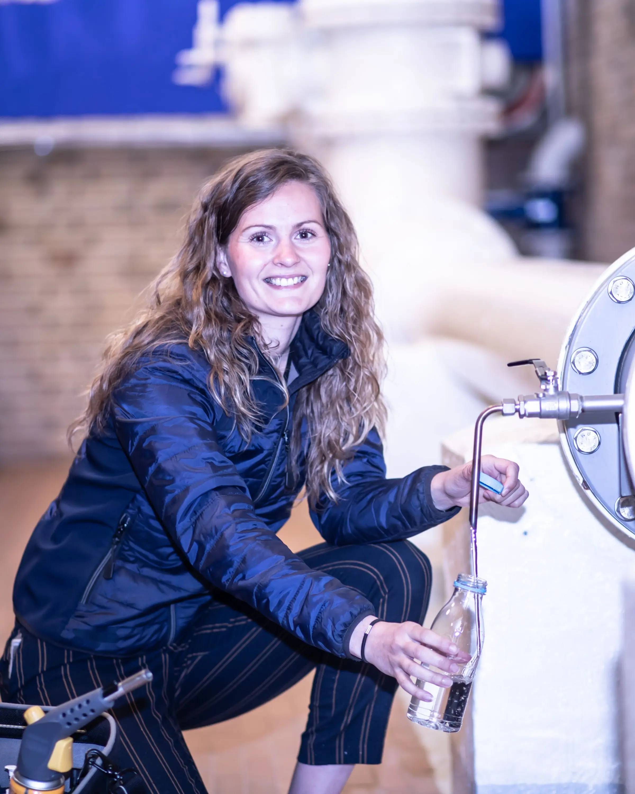 Mareike Kjellsen in dunkelblauer Steppjacke hockt an einer Wasserleitung und füllt am Probenahmeventil eine Flasche