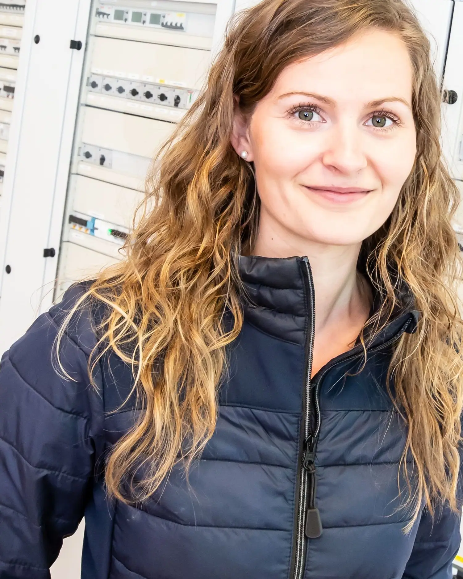 Mareike Kjellsen in dunkelblauer Steppjacke vor einer Reihe heller Schaltschränke