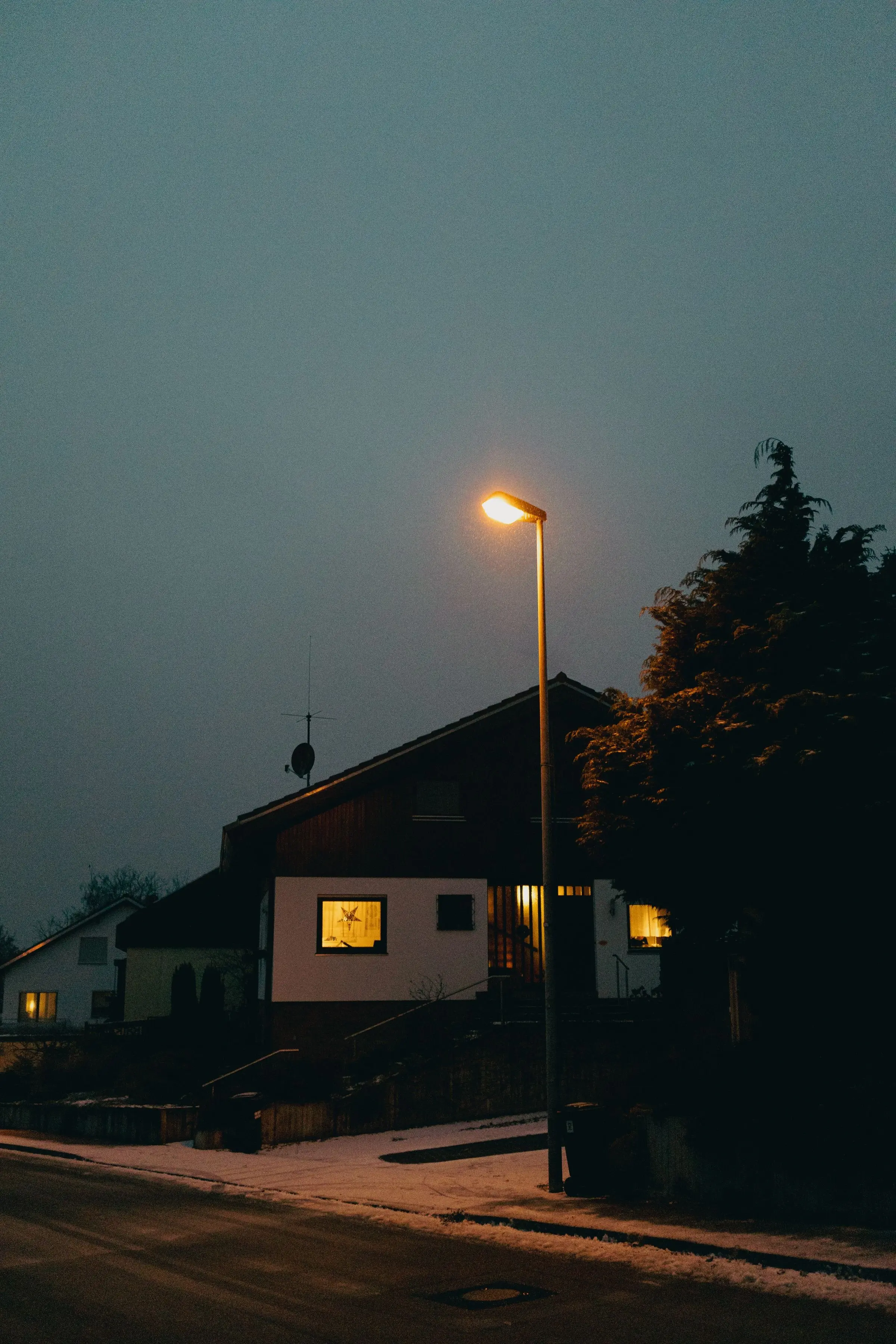 Dorfstraße im Winter bei Nacht, Schnee am Rand, eine Laterne und ein Haus mit erleuchtetem Fenster