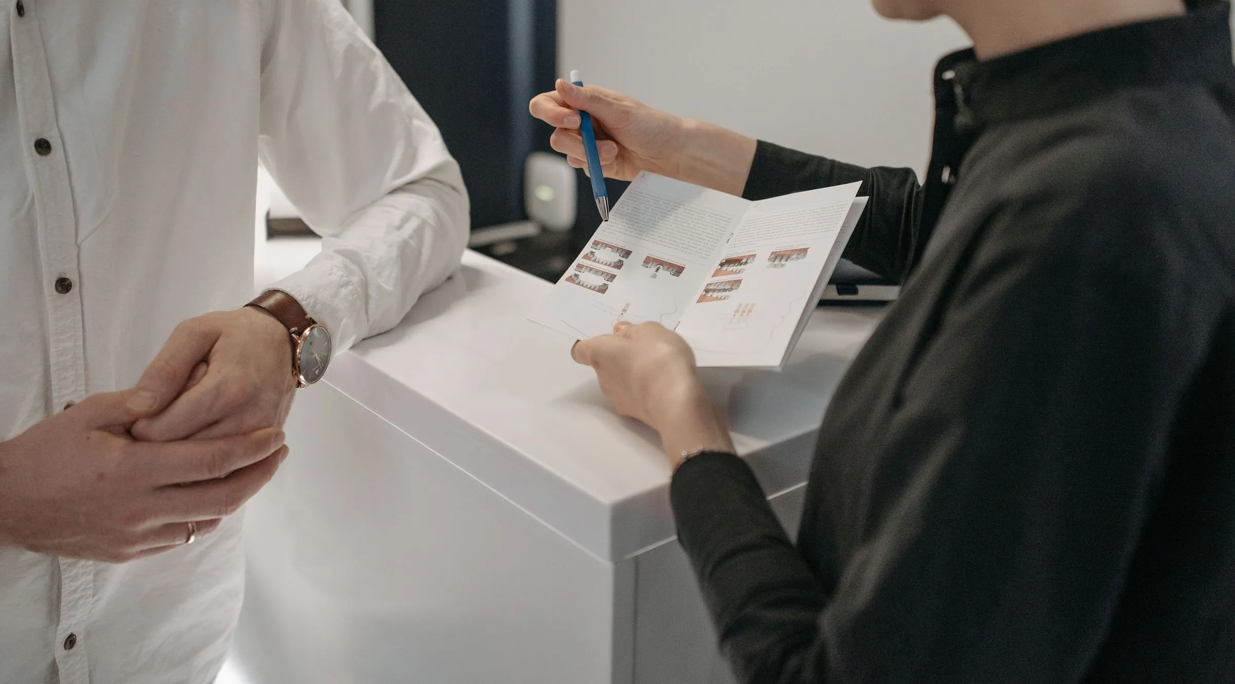 Am Empfangstresen zeigt eine Mitarbeiterin mit einem Stift auf eine aufgeklappte Broschüre, gegenüber steht ein Patient im weißen Hemd