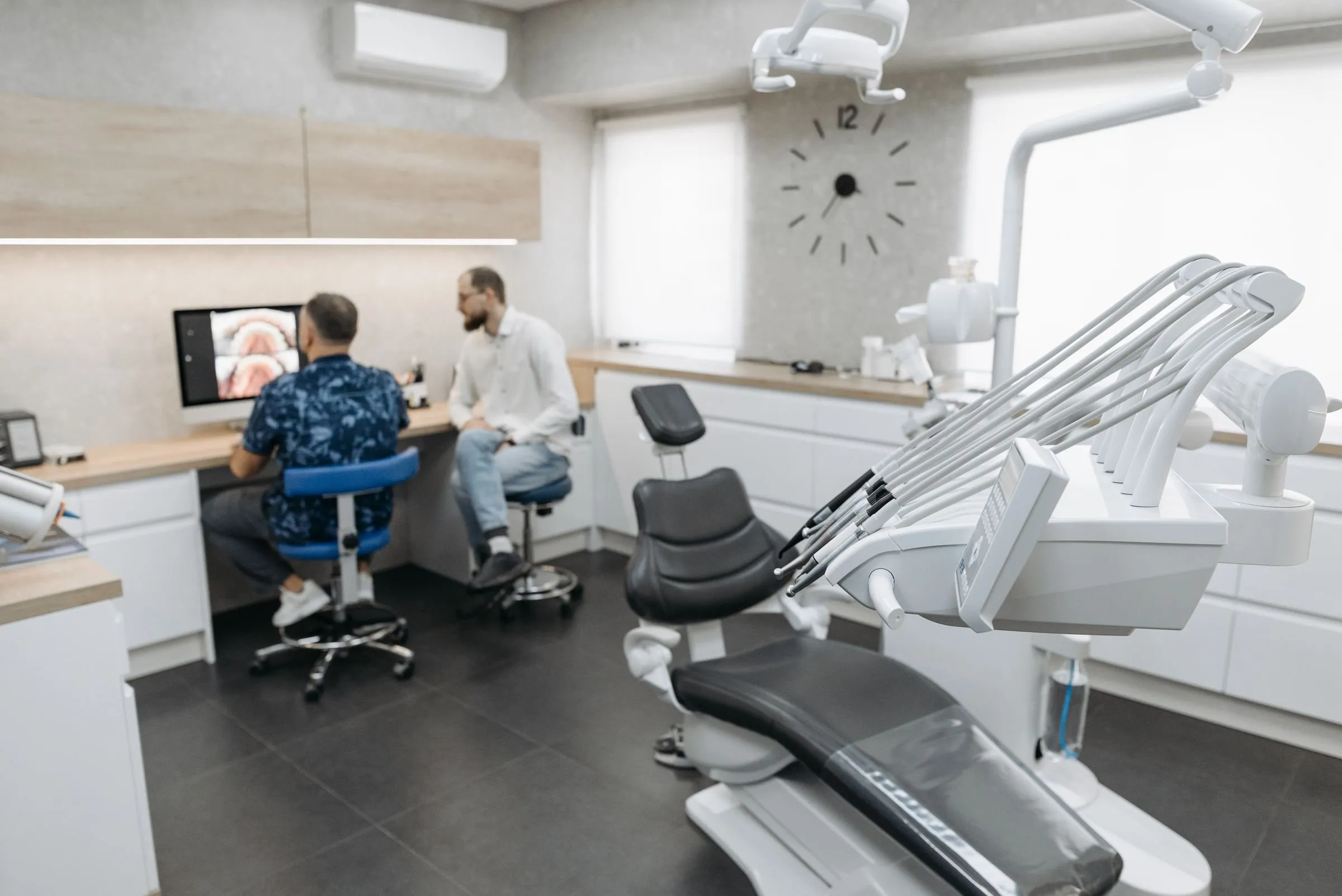Behandlungszimmer mit Fenster und großer Wanduhr; im Hintergrund sitzen Zahnarzt und Patient am Schreibtisch vor dem Bildschirm, vorne der Behandlungsstuhl