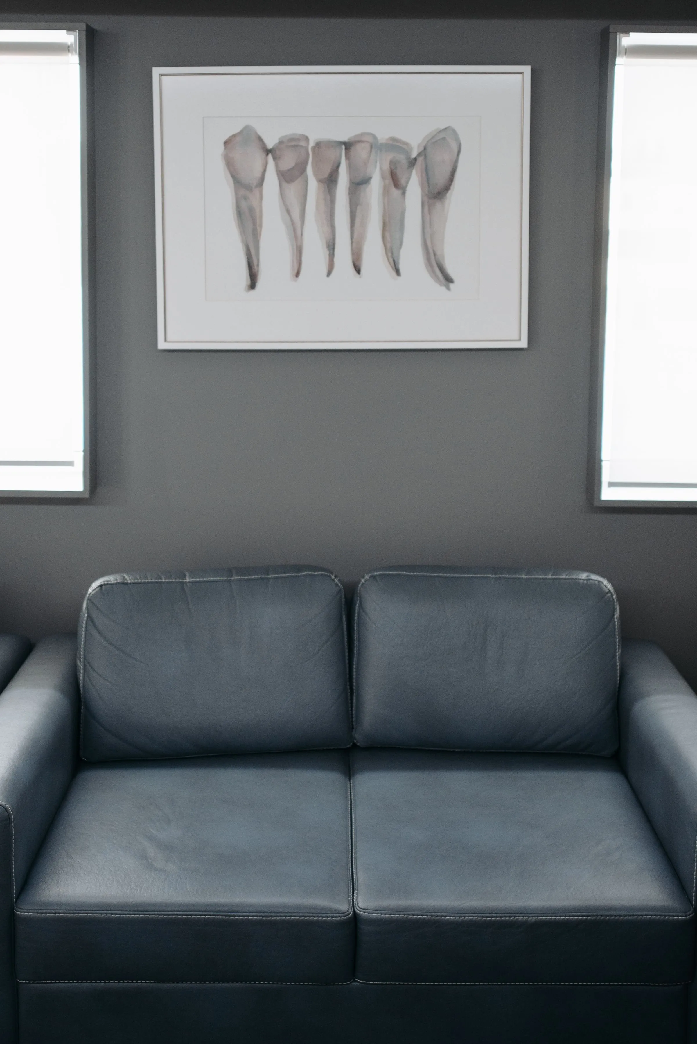 Wartebereich: graues Ledersofa vor einer dunkelgrauen Wand, darüber ein gerahmtes Aquarell mit Zahnwurzeln, links und rechts ein Fenster